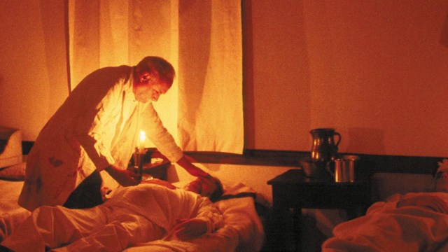 Surgeon in 1840s historic clothing treating a patient by candlelight in a historic hospital ward.