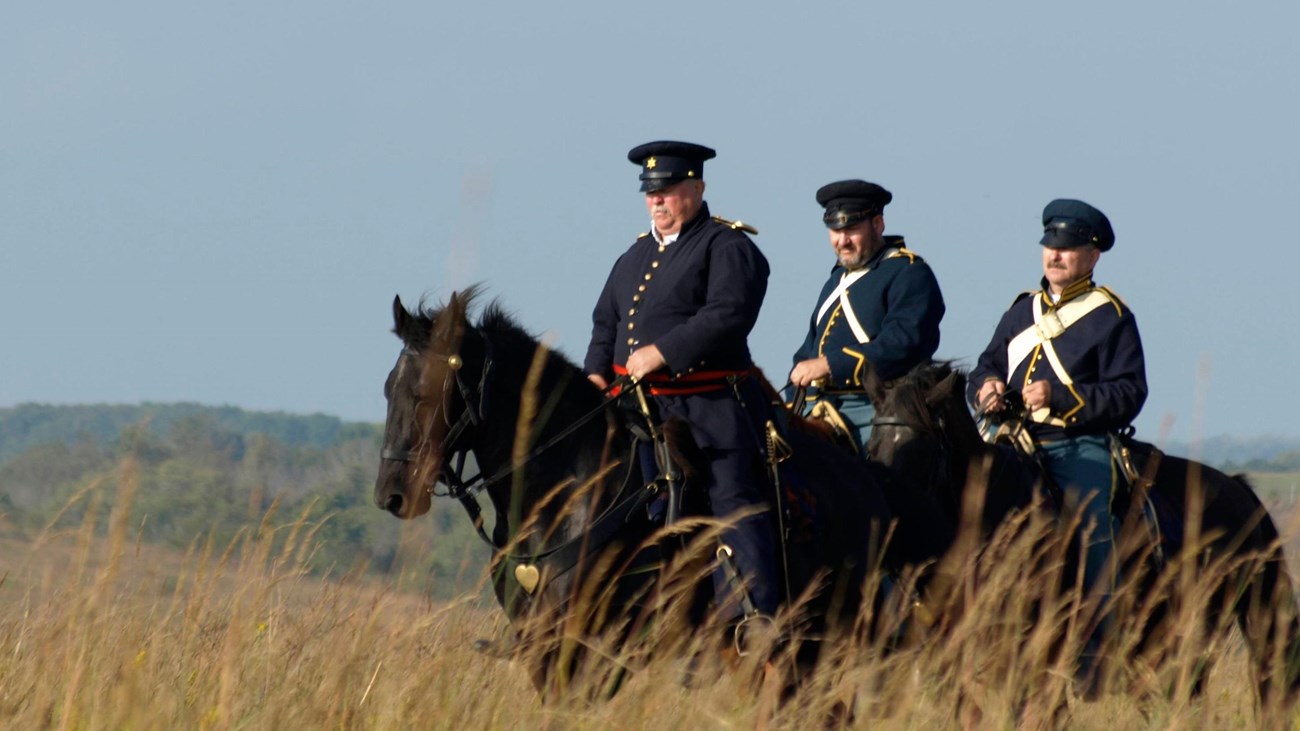 Three reenactors dressed in Dragoon Uniforms and on horseback riding through a tallgrass prairie.