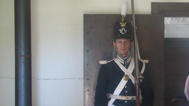 Soldier in infantry dress uniform wearing shako and holding a musket standing in front of a doorway.