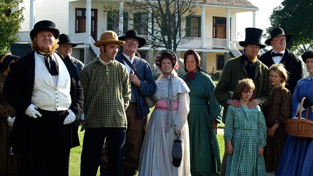 Men and women dressed in 1850s clothing on the grass with the white building in the background.