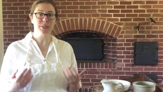 Woman in white clothing at a table with ceramic bowl, pitcher, and crock in front of a brick oven.