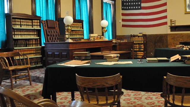 Bookshelves on both sides of a highbacked chair desk on left center of empty courtroom.