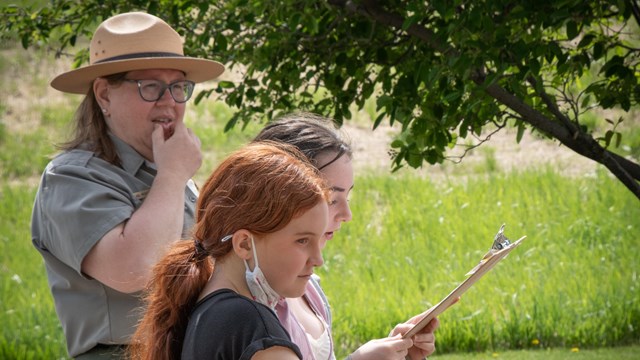 Two students stand with a park ranger. One student holds a cannon ball. 