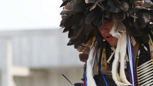 Profile portrait of man, wearing feather headdress, holding feather fan.