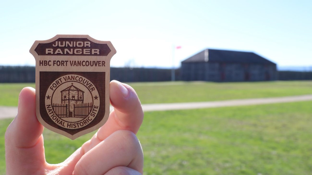 A hand holding up a Fort Vancouver junior ranger badge outdoors at the fort.
