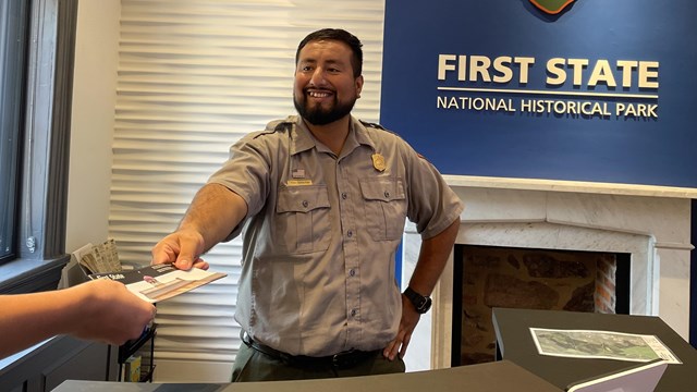 A male Park Ranger standing behind a front desk hands out a brochure.