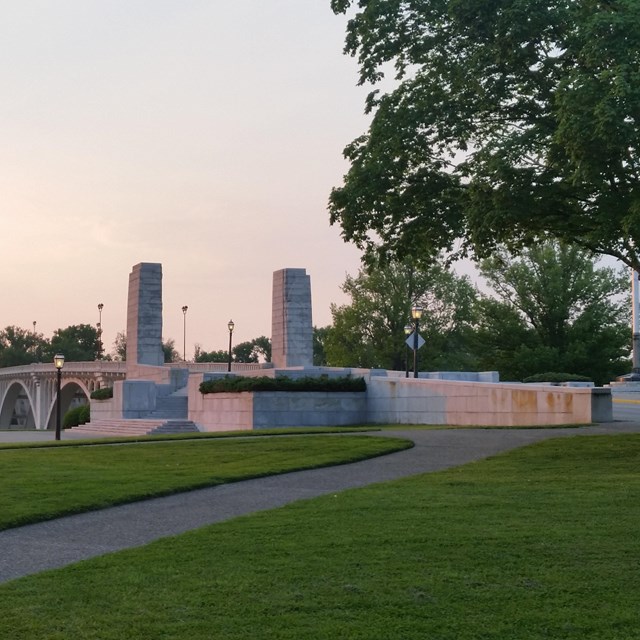 two gray rectangular columns rise above the roadway at the entrance to an arched concrete bridge.