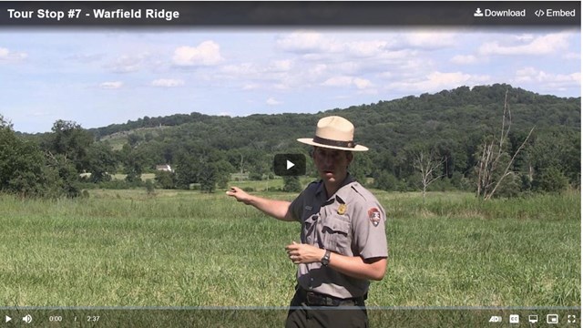 A park ranger points across a green field to two hills in the background