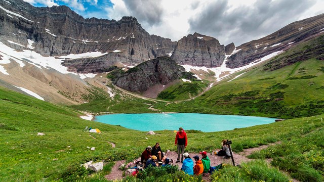 Visitors prepare a meal at a lakeside wilderness campsite near a mountain.