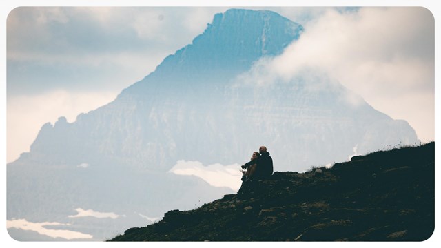 A small group of people sit on a hill with a mountain peak in the background. 