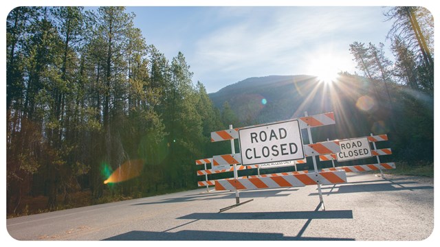 A sign says road closed and sun flares behind. 