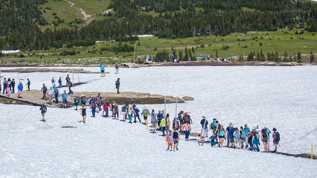 Crowds of hikers walk across a snowy meadow in the mountains. 