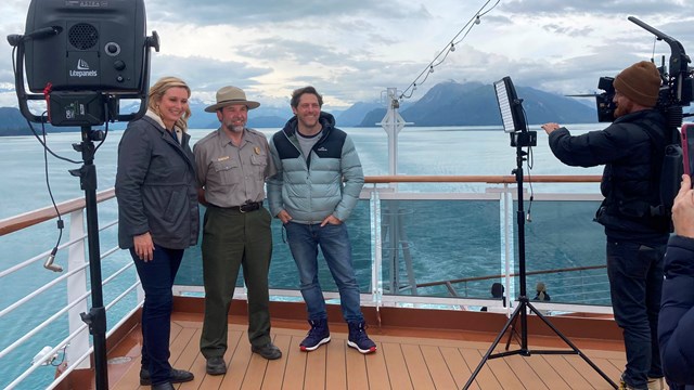 A park ranger stands between two people with camera crew filming and two lights illuminating them.