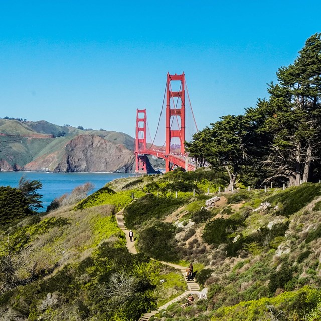 Trails along the bluffs with the a large red suspension bridge crossing a strait in the distance