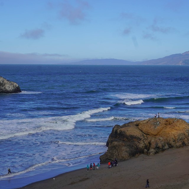 People walk, climb, and fish on a beach with large rocks and cliffs as waves crash on the shore.