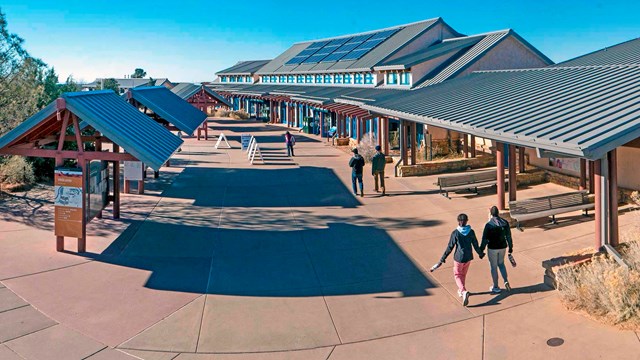 People walking across a plaza with information kiosks on the left and a long building on the right.