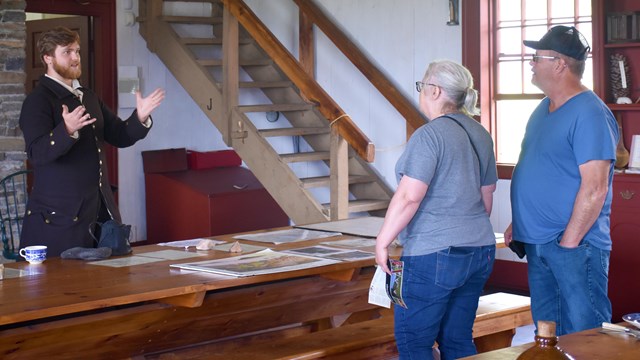 A person wearing historic clothing with hands in the air, speaking with two visitors indoors.