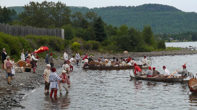 A brigade (several) canoes with people at the edge of a lake.