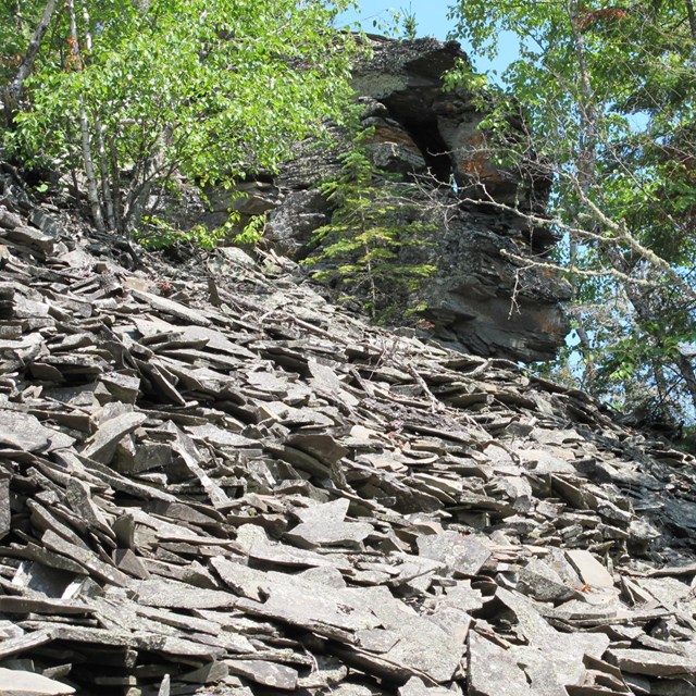 Broken gray slabs of rock in front of a cliff and green trees.