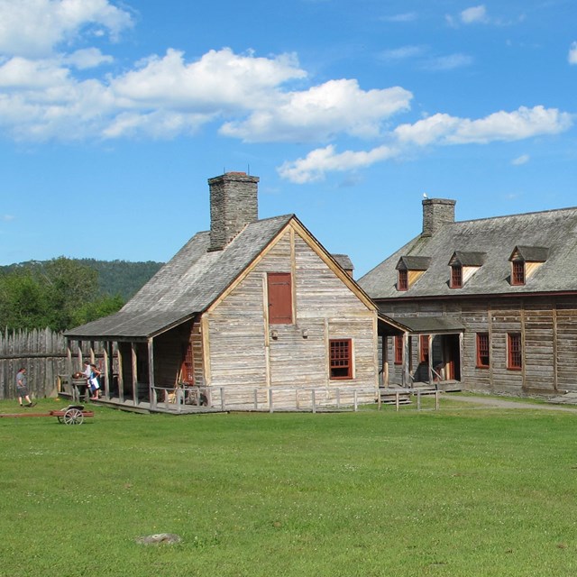 Reconstructed historic wood buildings on a green lawn, fenced by a palisade, in front of a bay.