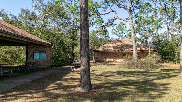 A brick pavilion and restroom building stand on concrete amongst grass and trees.