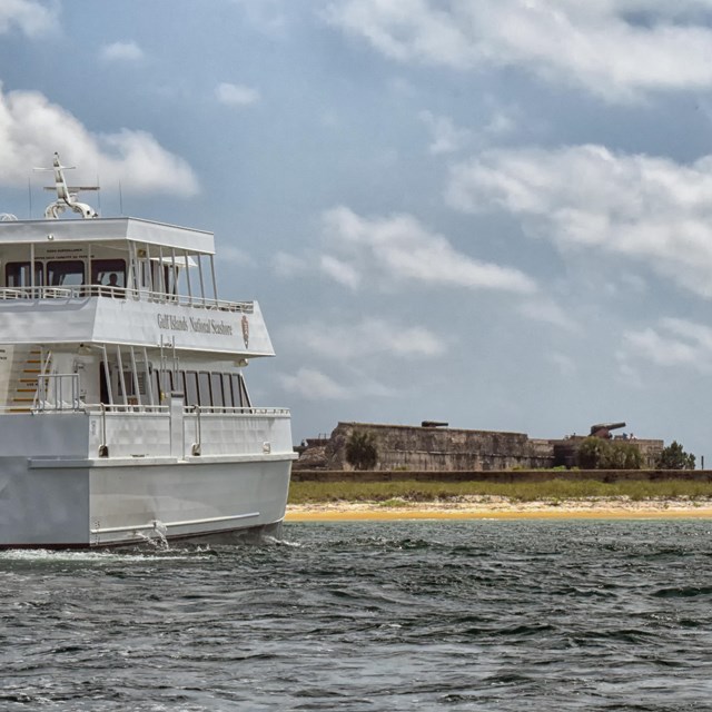 A large white ferry ships sits just off shore from a large masonry fort.