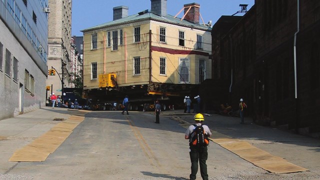 The Grange, elevated on hydraulics, is wheeled down 141st street as a man stands in front of it.