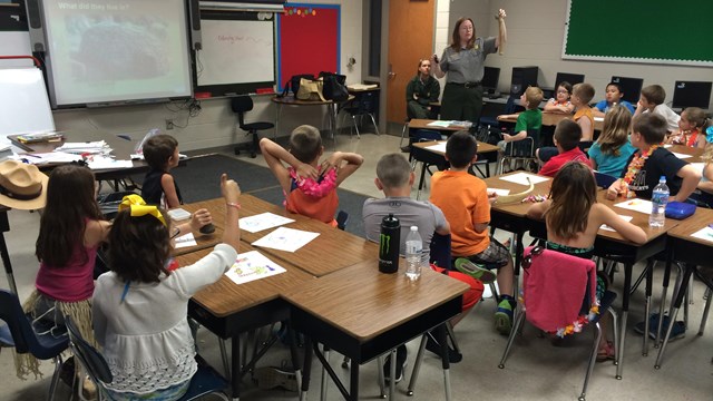 A group of students at their desks listen to a ranger as she holds a prehistoric tool.