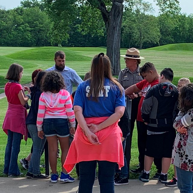 A group of students listen to a ranger on a patio in front of green grass and mounds