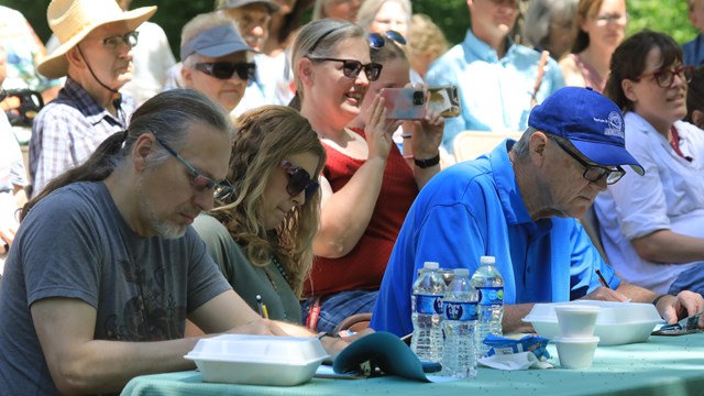 Three judges sit behind a table, writing on paper. A crowd behind them watches the stage.