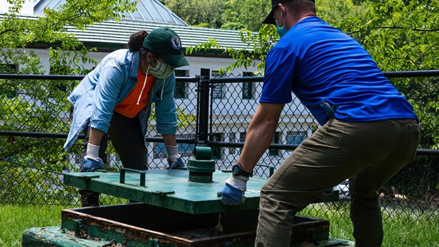 A woman and man work together to lift a large green metal lid off one of the spring boxes.