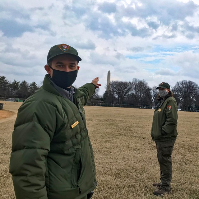 Two rangers in face masks, one pointing to the Washington Monument