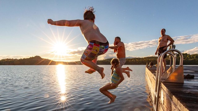 Two people jump off of a dock into calm waters as the sun sets in the background.