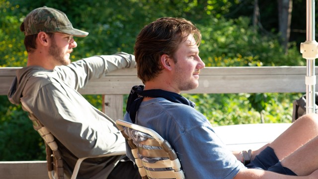 Two people sit in lawn chairs on a deck and look into the distance with green foliage behind them.