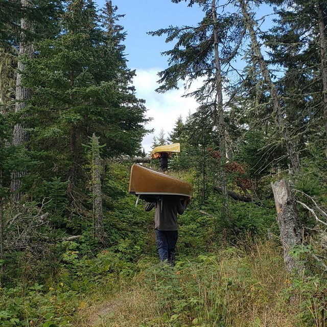 Two people, each portaging a canoe, hike on a trail up a hill surrounded by forest. 