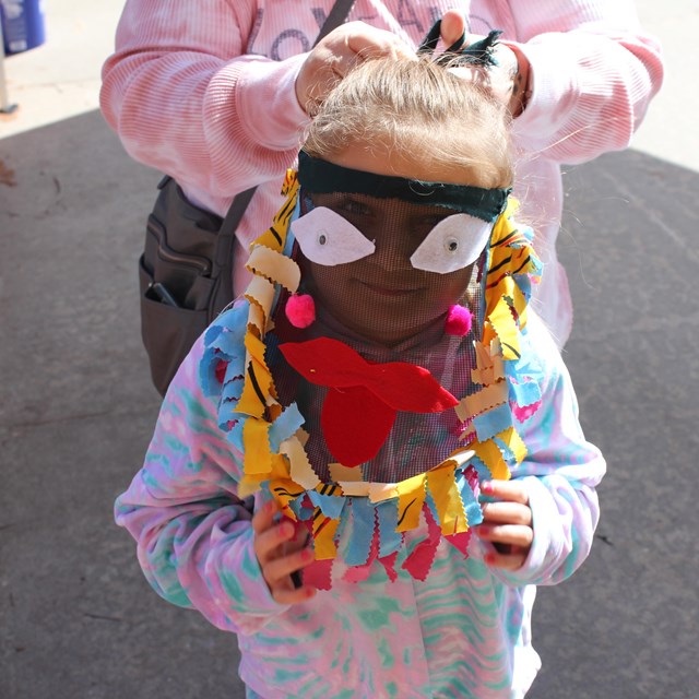 Outside daytime. Young girl smiles behind a mesh mask with felt on it.