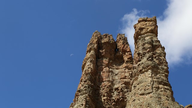 Stone pillars reach into a blue sky with the pale outline of the moon visible to the left.