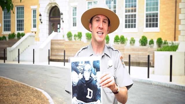 A park ranger stands in front of a yellow bricked building holding a photograph of a young JFK 