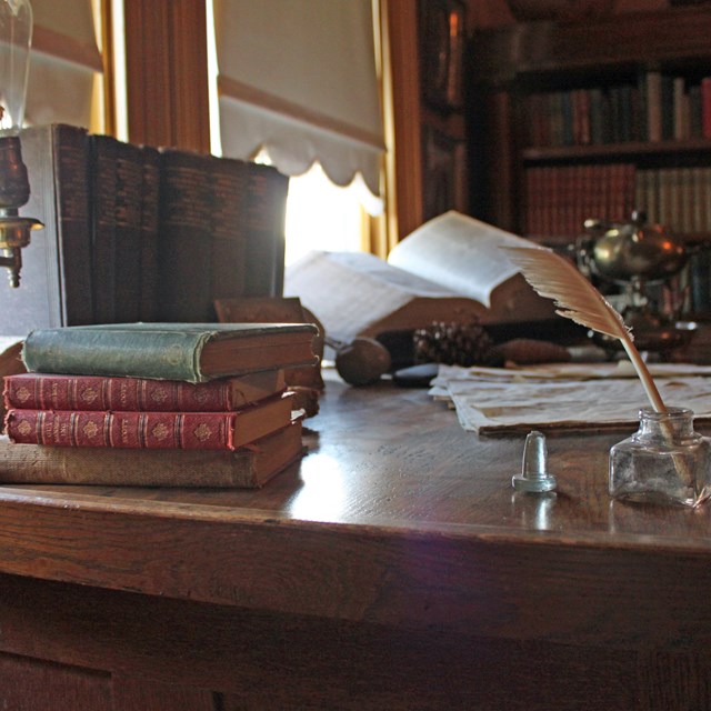 A desk is seen with piles of books and various items. Light shines through a window. 