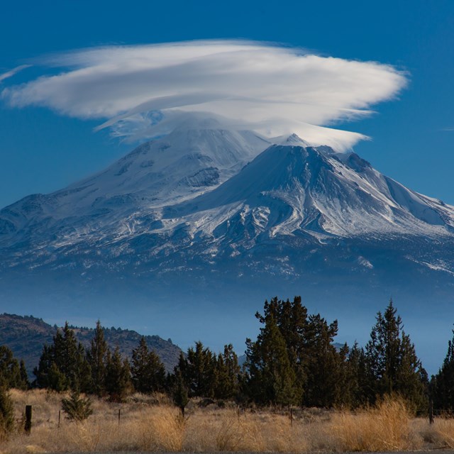 A majestic mountain piercing a cloud. Trees and a plane in the forground. 