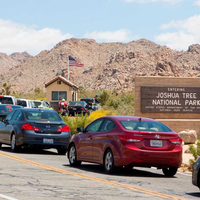 A line of cars on a road passing a sign that reads Entering Joshua Tree National Park