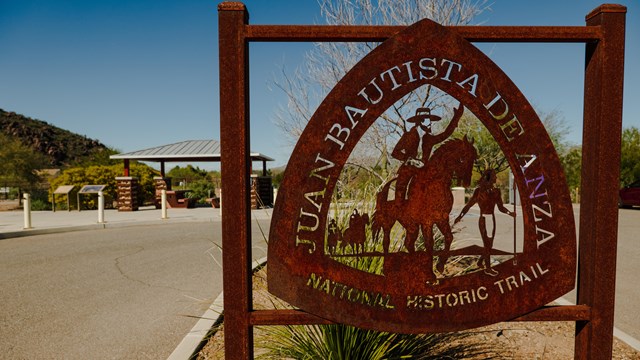 A rusted metal sign in front of a desert park shows the Anza Trail National Historic Trail emblem