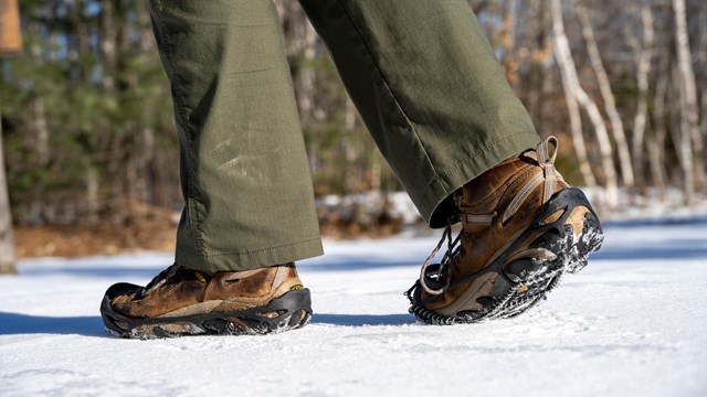 A pair of feet in brown boots with snow spikes walks on snowy ground.