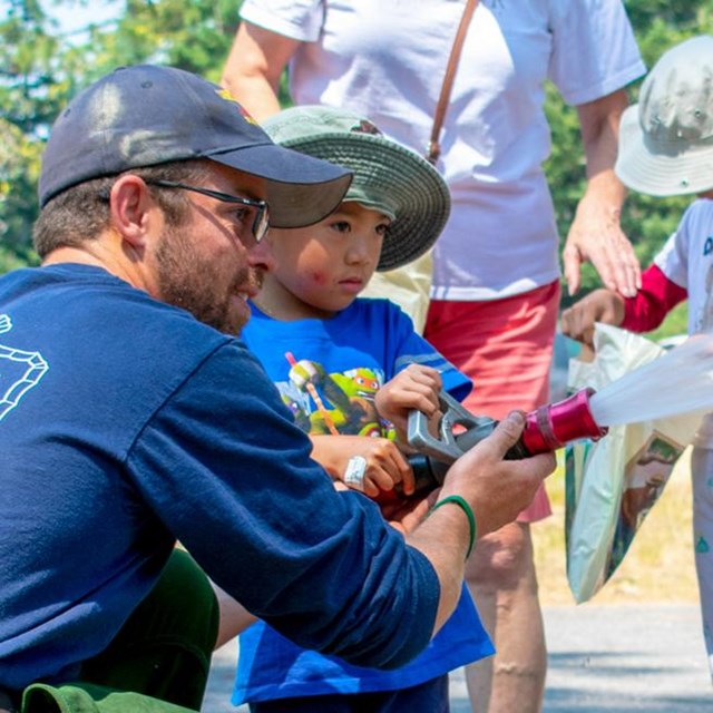 A firefighter helps a young boy use a fire hose at a public demonstration.