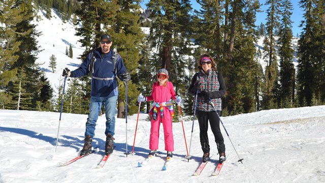 A man, woman, and young girl on cross-country skis in a mountain landscape.