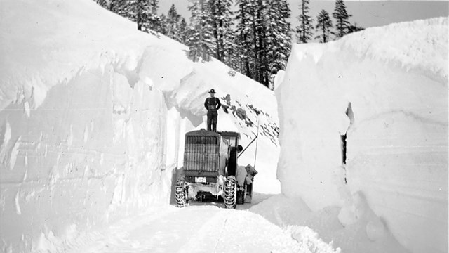 A man stands on a snowplow flanked by walls of snow