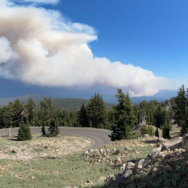 A road winds down a mountain slope with smoke rising from a wildfire in the distance. 