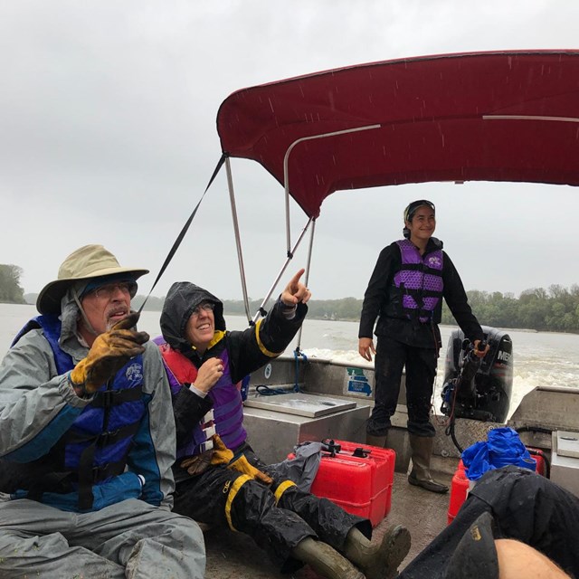 Young person operates engine on open boat in rain. Two people point into the distance