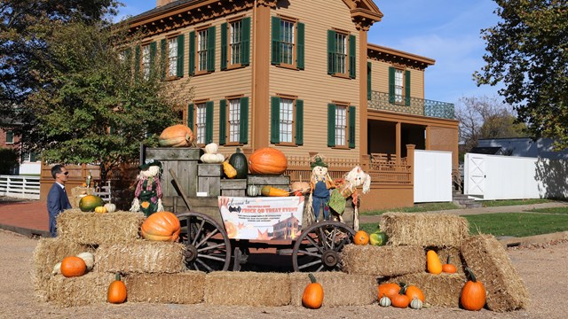 A two story tan house with green shutters. In front a wagon with pumpkins