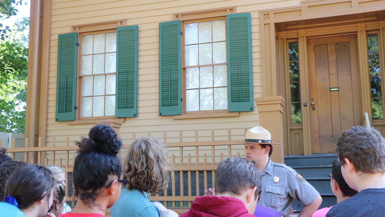 A ranger stands in front of a two story tan house with a group of people